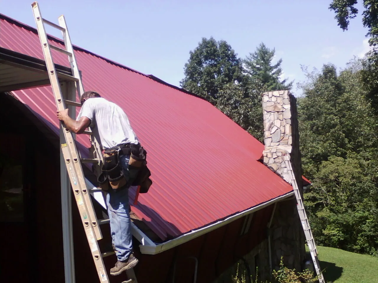 Roofer on a ladder beside a red metal roof and stone chimney — Soffit & Fascia Repair by Roofing Brothers in Canova